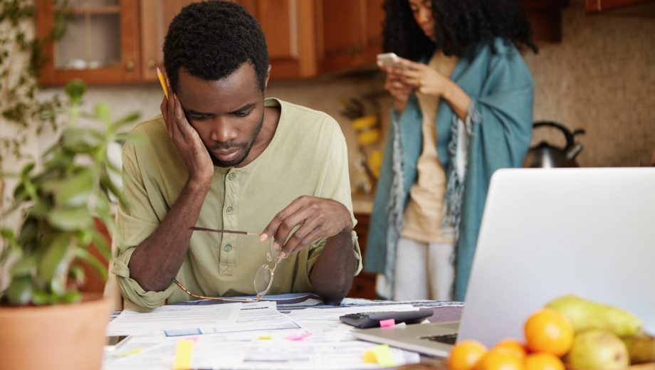 No money. Young family in financial trouble. Frustrated African man with glasses and pencil in his hands, looking at papers on table with stressed and puzzled expression, paying bills online at home